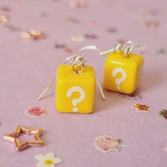 two yellow square earrings with question marks on them sitting on a purple table covered in confetti