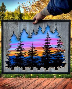 a person holding up a piece of artwork with trees in the background and sunset behind them