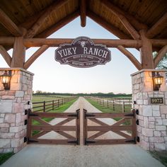 The Yuky Ranch entrance features a wooden arch with a vintage-style metal sign, supported by stone columns with lanterns for lighting. The double wooden gate, secured with metal hinges, opens to a gravel driveway bordered by wooden fences, blending durability with rustic charm. Ranch Gate Entrance, Ranch Driveway Entrance, Farm Driveway Entrance, Farm Entrance Ideas Driveways, Ranch Gates Entrance Ideas, Gated Community Entrance, Ranch Signs Entrance, Gate Entrance Ideas