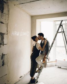 a man and woman standing on a ladder kissing in an unfinished room with graffiti written on the wall