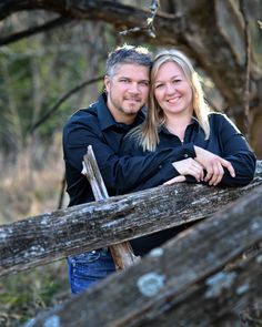 a man and woman standing next to each other in front of a wooden fence with trees behind them