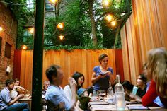 a group of people sitting around a table in a small outdoor dining area with lights hanging from the ceiling