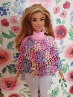 a barbie doll with blonde hair wearing a pink sweater and gray pants, standing in front of a floral wallpaper