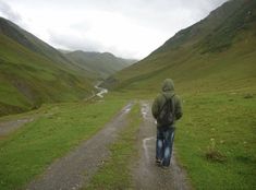 a man walking down a dirt road in the middle of a green valley with mountains behind him