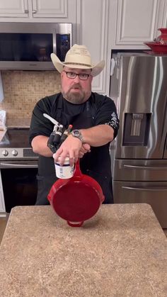 a man in a cowboy hat is pouring something into a red pot on a kitchen counter