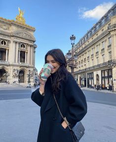 a woman standing in front of a building drinking from a cup while wearing a black coat