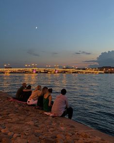 four people sitting on the edge of a river watching the sun set