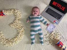 a baby laying on a bed next to a laptop computer and popcorn buckets with the word netflix spelled out