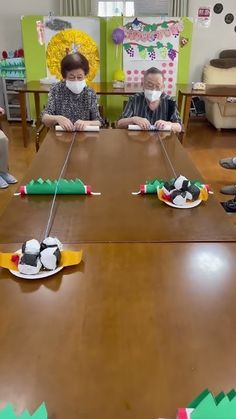 several people sitting at a long table with masks on their faces and paper boats in front of them