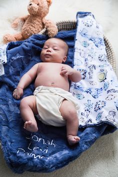 a baby laying on top of a blue blanket next to a teddy bear in a basket