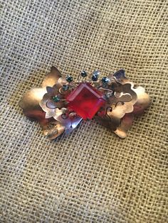 Here is a fantastic, three dimensional brooch which is probably from the 1940's. It is in the shape of a bow-tie with scalloped edges and pierced top layer. The center has a prong-set, large, red glass stone. This stone is crowned with eight rhinestones which stand up on metal stalks. The stones are white, green, and blue. The piece is marked sterling on its back, and it has a copper-colored wash. It closes with a long pin on the back which fits into a "C"  with a roll-over clasp. The mechanism still functions well, although the pin is bent slightly. This is a large, three dimensional piece. It measures 4" by 2", and it stands 1" high. It is still in good vintage condition with some tarnish. It is a one-of-a-kind statement piece and a conversation starter. Definitely unique and exotic. Handmade Vintage Red Brooches, Vintage Red Brooches For Party, Vintage Red Pins For Party, Vintage Red Brooches For Formal Occasions, Silver Brooch Pin, Sterling Silver Brooch, Silver Brooch, Scalloped Edges, Red Glass