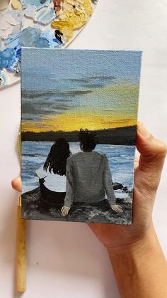 a person holding up a small painting with water and sky in the background, next to a paintbrush