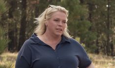 a woman in a blue shirt is standing near some pine trees and looking at the camera