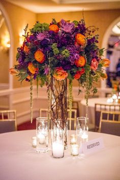 a tall vase filled with lots of purple and orange flowers next to some candles on top of a table