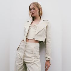 a woman with blonde hair standing in front of a white wall wearing beige pants and a jacket