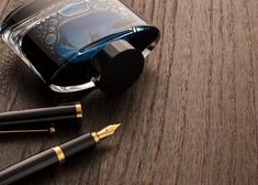 two fountain pens sitting on top of a wooden table next to a bottle of ink