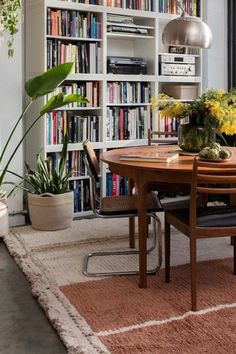 a dining room table surrounded by bookshelves and potted plants in the corner