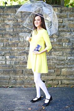 a woman in a yellow dress holding an umbrella and a book while standing next to a brick wall