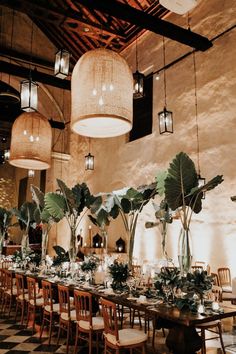 a long table with chairs and plants in vases on top of it, surrounded by hanging lights