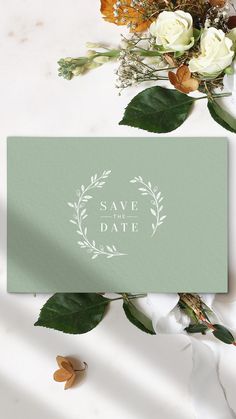 save the date card with white roses and greenery on a marble surface next to a bouquet of flowers