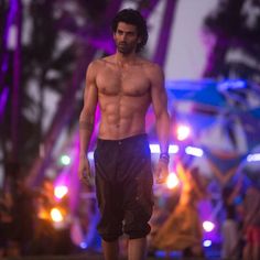 a shirtless man walks down the runway at an outdoor event in front of purple lights