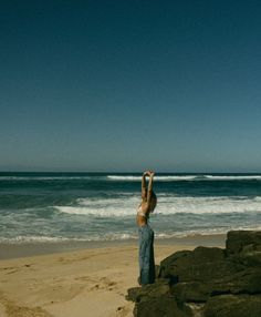 a woman standing on top of a sandy beach next to the ocean with her arms in the air