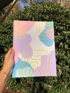 a hand holding up a piece of paper with an abstract design on it in front of some bushes