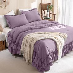 a bed with purple comforter and pillows in a room next to a white chair