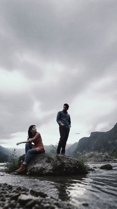 two people sitting on top of a rock in the middle of a river with mountains in the background