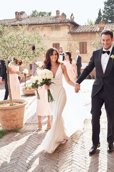 a bride and groom holding hands as they walk down the street with their wedding party in the background