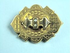 "Very pretty 18k gold pin from the mid Victorian Period c. 1870 is formed of a hollow shield-shaped body etched with a wavy pattern, tiny star bursts and decorative trim.  The top has a stylized ribbon surmounted that is decorated with taille d'Epergne enameling and is set with two grey seed pearls.  This is a fine example of the etching that was typical of the period.  The back has a C-clasp and tube hinge and also has a hole that likely held a hook or loop to have it serve as a pendant.  Measu Ornate Engraved Yellow Gold Brooches, Ornate Gold Locket Brooches, Victorian Gold Engraved Brooches, Victorian Engraved Gold Brooches, Victorian Engraved Medallion Brooches, Ornate Gold Engraved Brooches, Victorian Gold Brooch With Screw Back, Wavy Pattern, Gold Pin