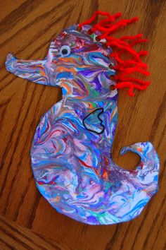 a paper plate seahorse craft with red hair on the head and tail, sitting on top of a wooden table