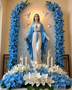 a statue of the virgin mary surrounded by blue and white flowers with candles in front of it