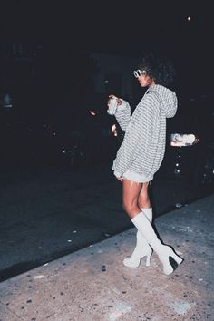 a woman standing on the sidewalk at night wearing white boots and a sweater over her shoulders