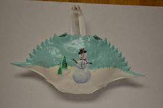 a paper plate shaped like a snowman with trees in the background and an ornament hanging from it