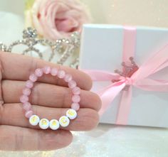 a hand holding a pink bracelet with white beads and yellow letters on it next to a gift box