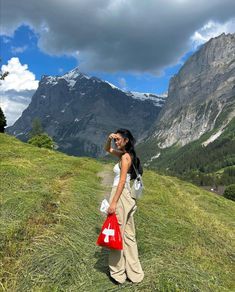 a woman standing on top of a grass covered hillside holding a red bag and talking on a cell phone