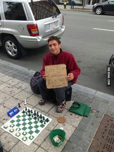 a man is sitting on the sidewalk holding a sign that says, let's play chess