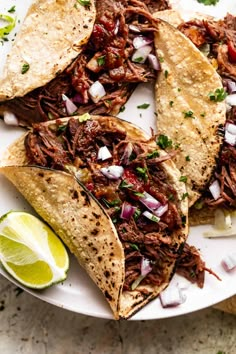 three shredded beef tacos on a white plate with lime wedges and cilantro