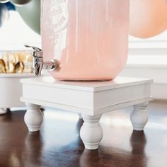 a pink beverage dispenser sitting on top of a wooden table with balloons in the background