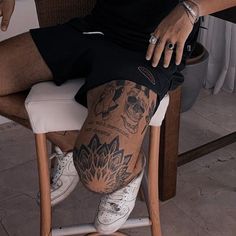 a man with tattoos sitting on a chair in front of a table and holding his leg up