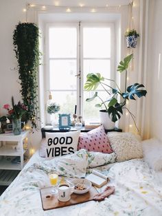 a bed with white sheets and pillows in front of a window filled with potted plants