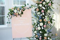 a pink sign with flowers on it next to a tall flower arrangement in front of a window