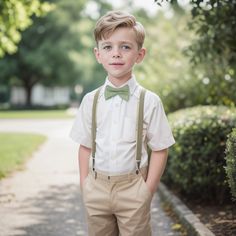 a young boy wearing suspenders and a bow tie standing on a sidewalk in front of bushes