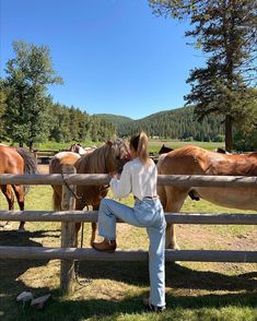 a woman is sitting on a fence with horses in the field behind her and looking at them