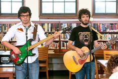 two young men are playing guitars in front of microphones and bookshelves, while one man is singing