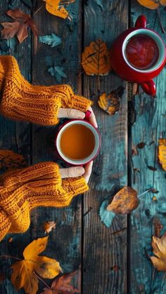 two hands in mitts holding cups of tea on a wooden table with autumn leaves