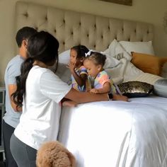 a group of people standing around a small child on a bed in a room with white sheets and pillows