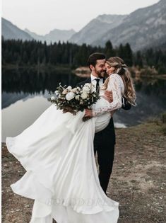 a bride and groom standing next to each other in front of a lake with mountains