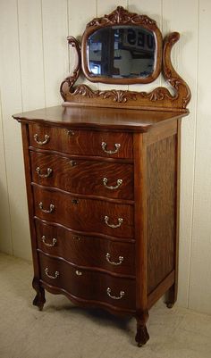 an ornate wooden dresser with a mirror on it's top and bottom drawer, in front of a white wall